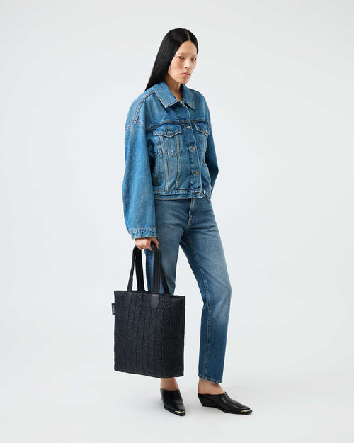 Model wearing a blue denim jacket and jeans holds a black quilted Vee Shopper tote.