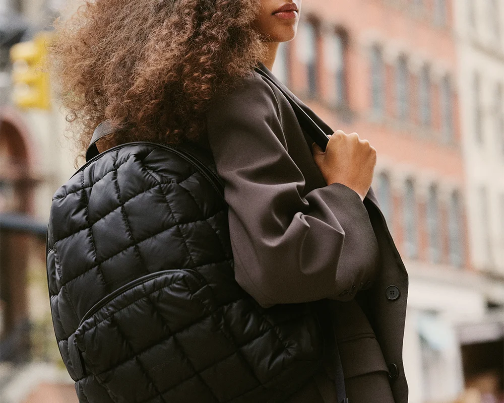 Woman wearing a Vee Collective Porter backpack in matt black over one shoulder, upper body only, with a blurred urban background.